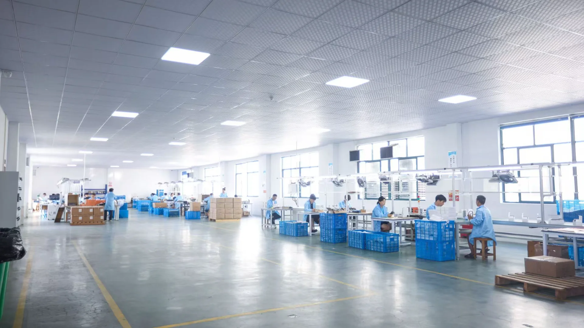 Spacious modern factory floor with workers at assembly stations, wearing blue uniforms, surrounded by blue bins and boxes.