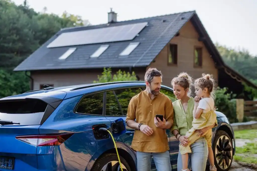 Family standing near an electric vehicle being charged outside their home, equipped with solar panels on the roof.