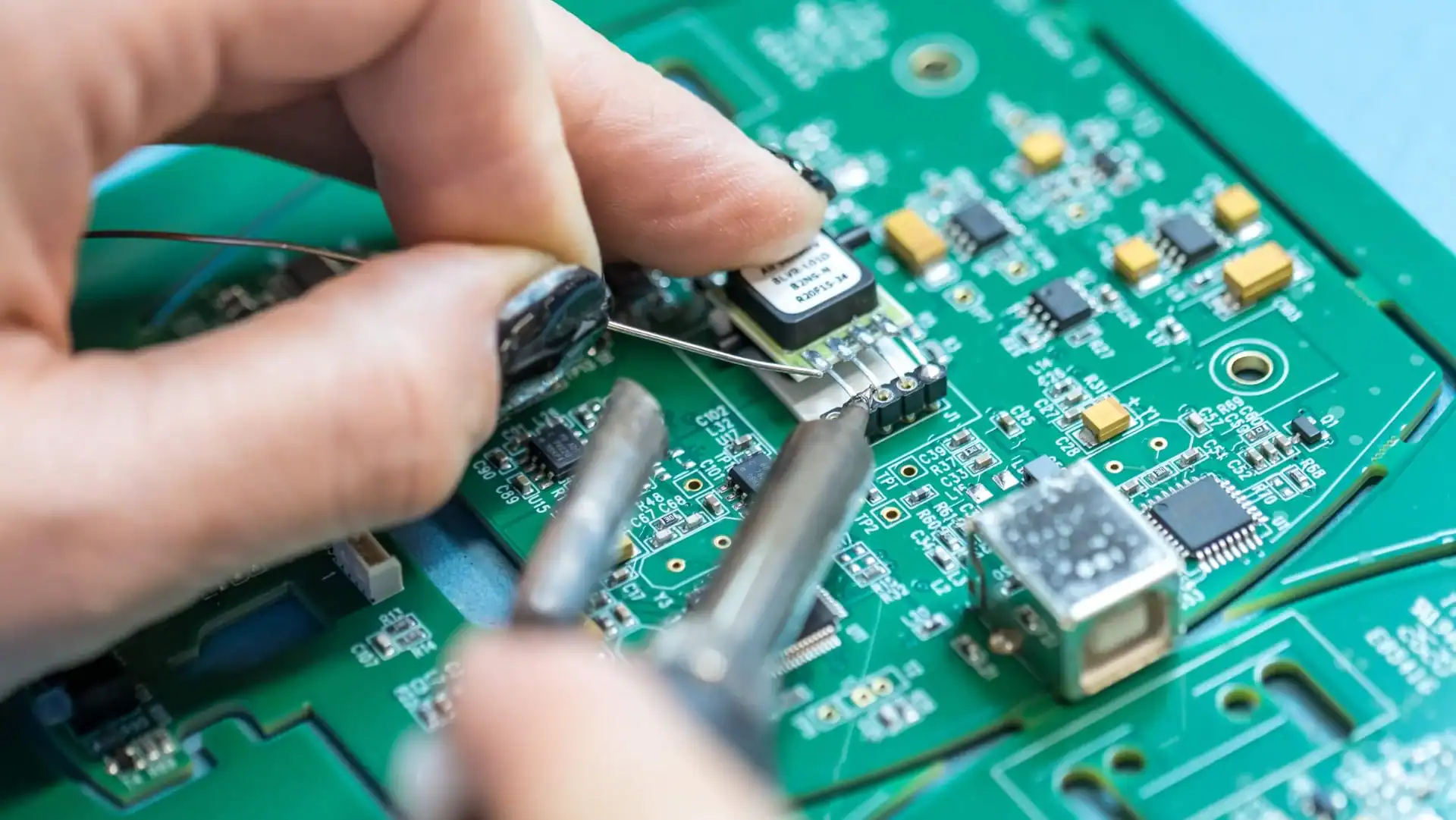 복잡한 회로와 도구로 녹색 회로 기판에 전자 부품을 납땜하는 손을 클로즈업합니다.