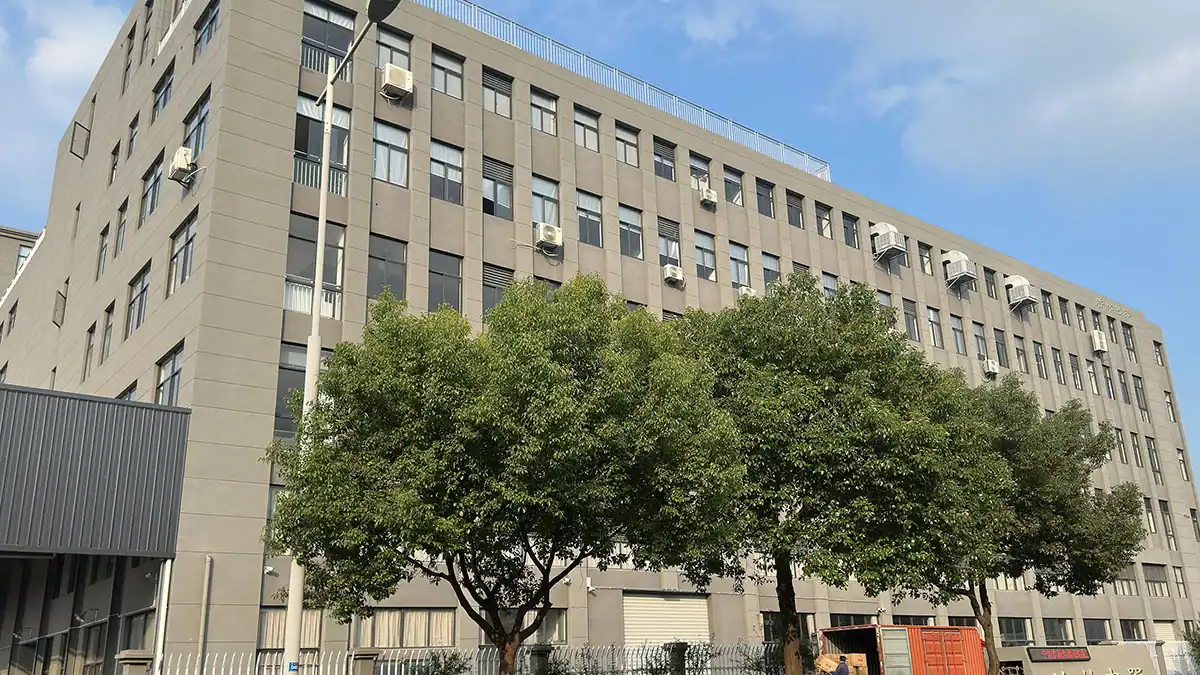 A multi-story industrial building with numerous windows and air conditioning units, surrounded by lush trees under a sunny blue sky.