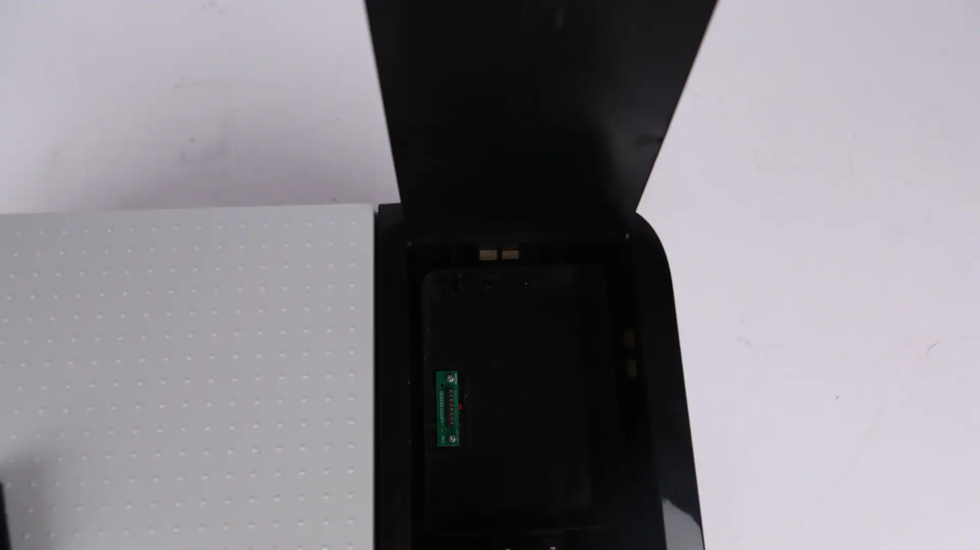 Open black compartment of a device with a visible circuit inlay, placed on a white surface with a patterned grey panel.