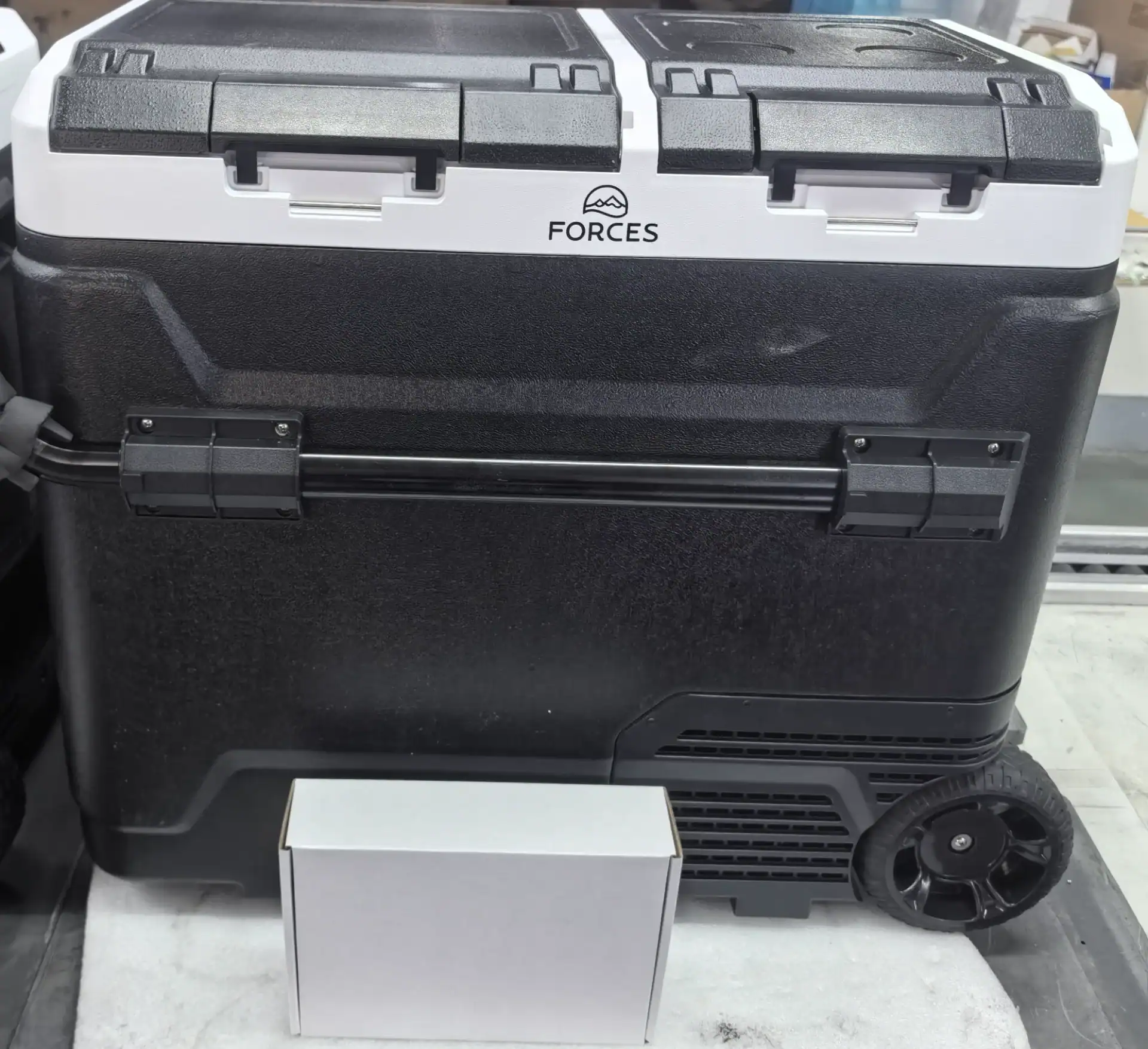 Black and white portable car fridge with wheels, handle, and dual lid design, placed on a surface with a small white box in front.