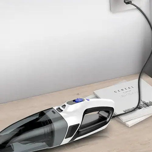 Compact portable handheld vacuum cleaner in white and black placed on a desk near a power outlet, charging with a cable.