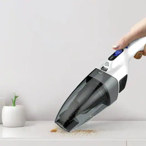 Hand using a portable handheld vacuum cleaner to clean dirt and crumbs from a white surface, with a small decorative plant nearby.