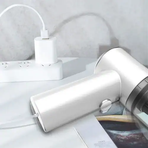 Compact white portable vacuum cleaner charging on a desk, connected to a power outlet via a USB cable, placed next to a power strip and open magazine.
